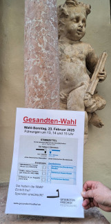 Ein Wahlzettel mit der Aufschrift Gesandten-Wahl vor einem Puto an einer Säule (Teil eines Grabmals auf dem Gesandtenfriedhof). Auf dem Stimmzettel sind die Namen von Gesandten zur Wahl gestellt.